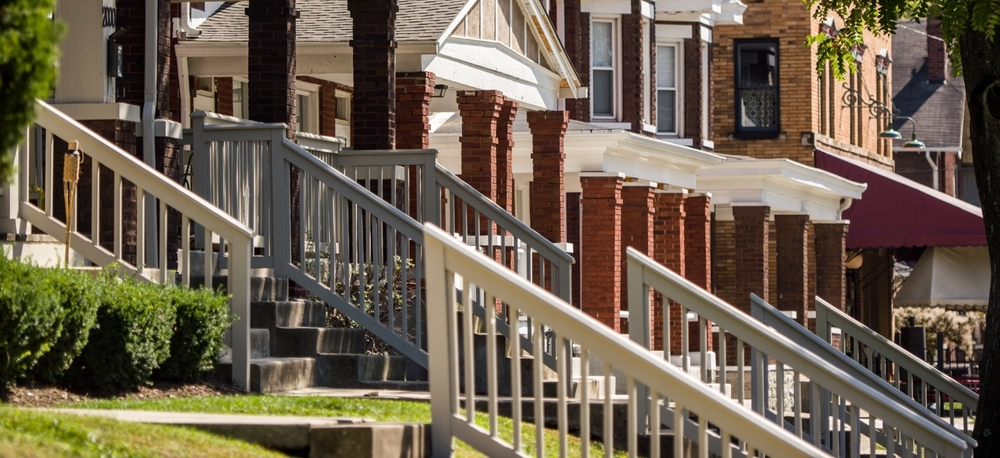 A tree-lined street of historic early-1900s brick homes in Columbus, Ohio, with white painted front porch staircases and railings repeating into the distance.